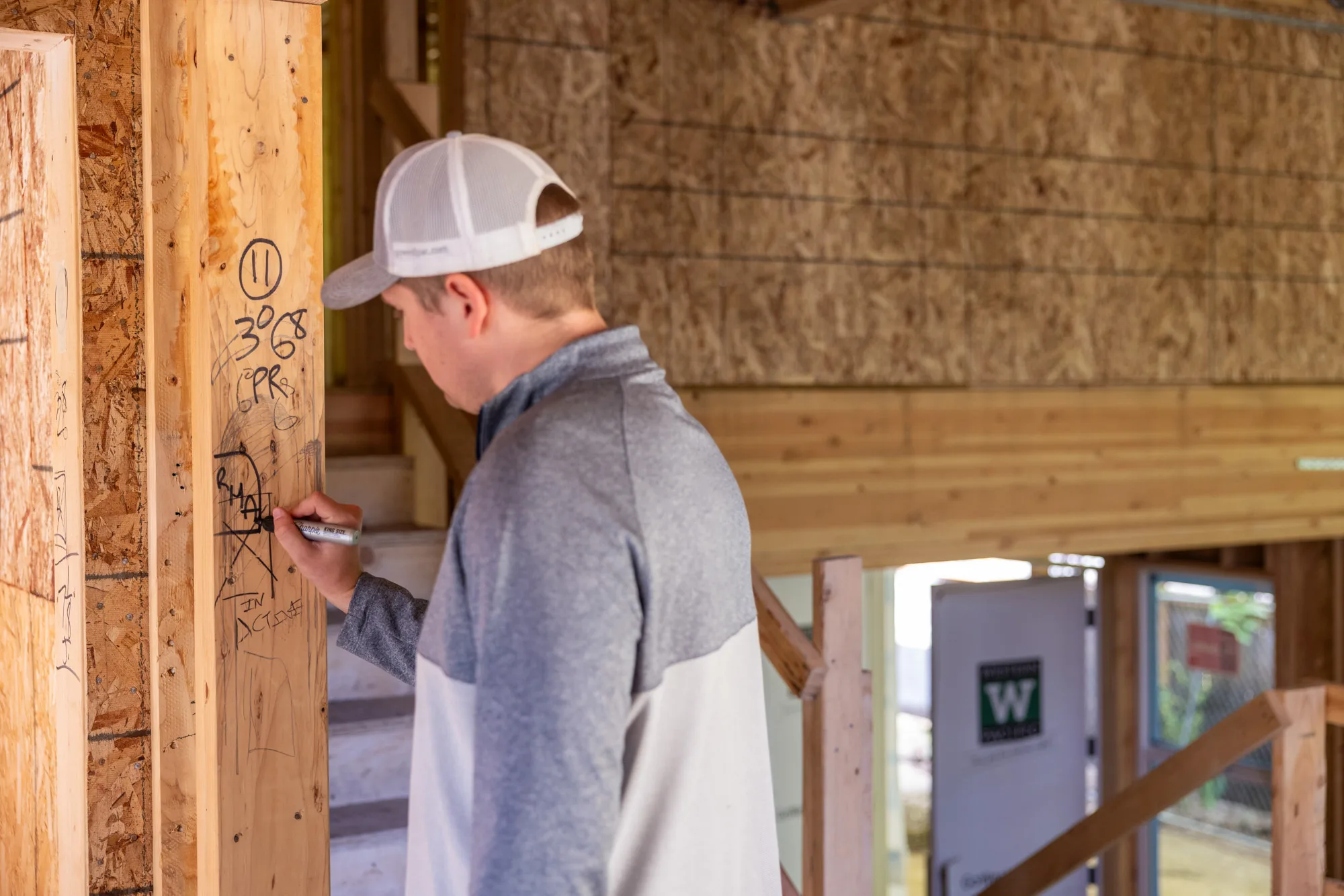 Person wearing a gray and white jacket and light gray baseball cap writing on a wooden beam inside a building under construction.