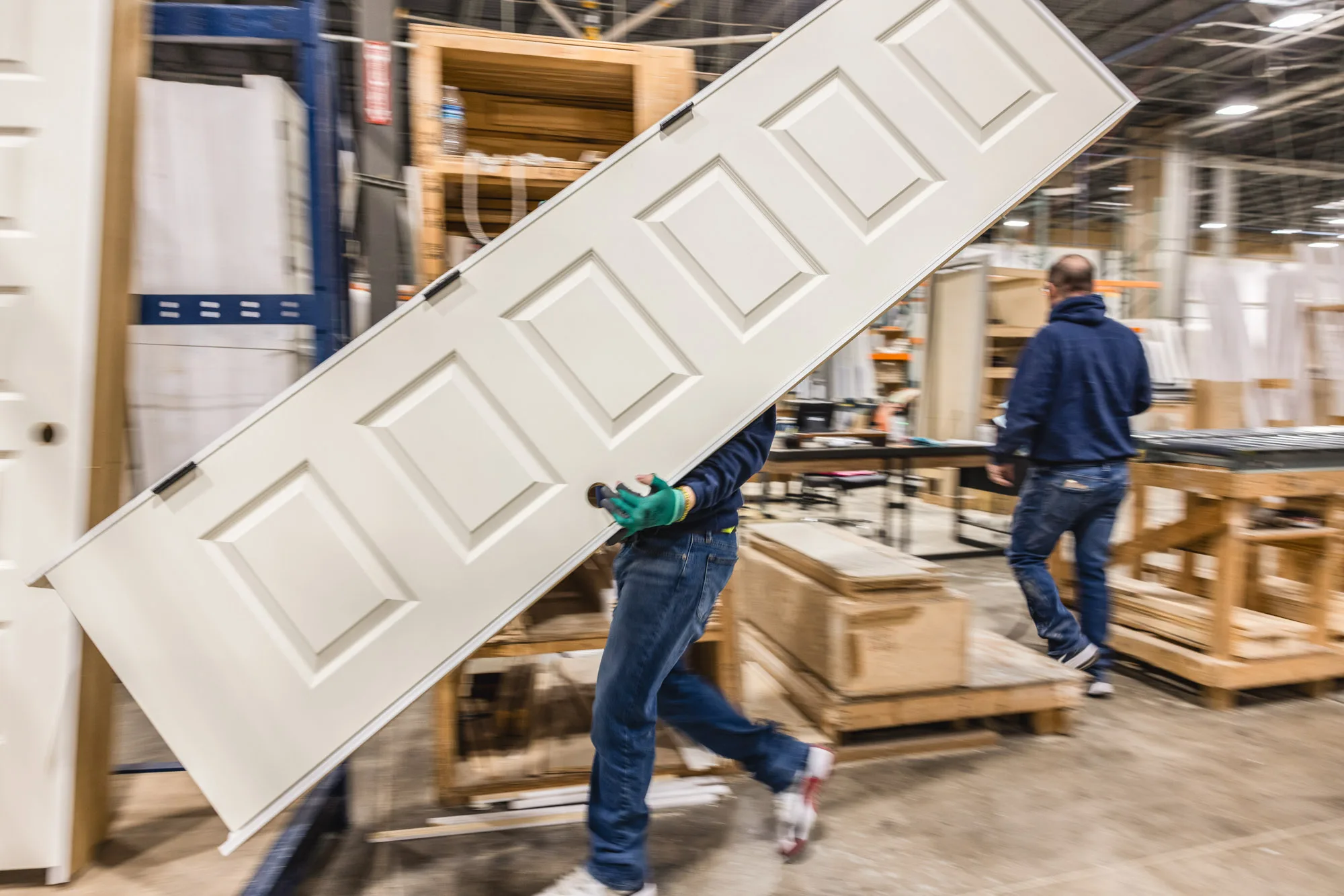 Worker in a warehouse carrying a large white panel door, with another person walking away in the background among shelves and pallets of materials.