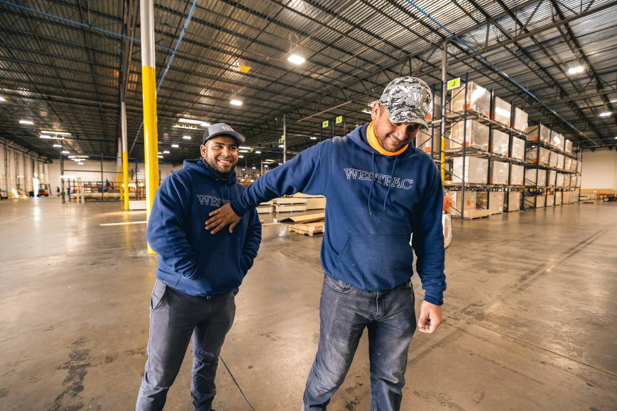 Two warehouse workers smiling and interacting inside a large storage facility, wearing navy blue hoodies with the word WESTPAC and caps, with shelves of stacked pallets in the background.