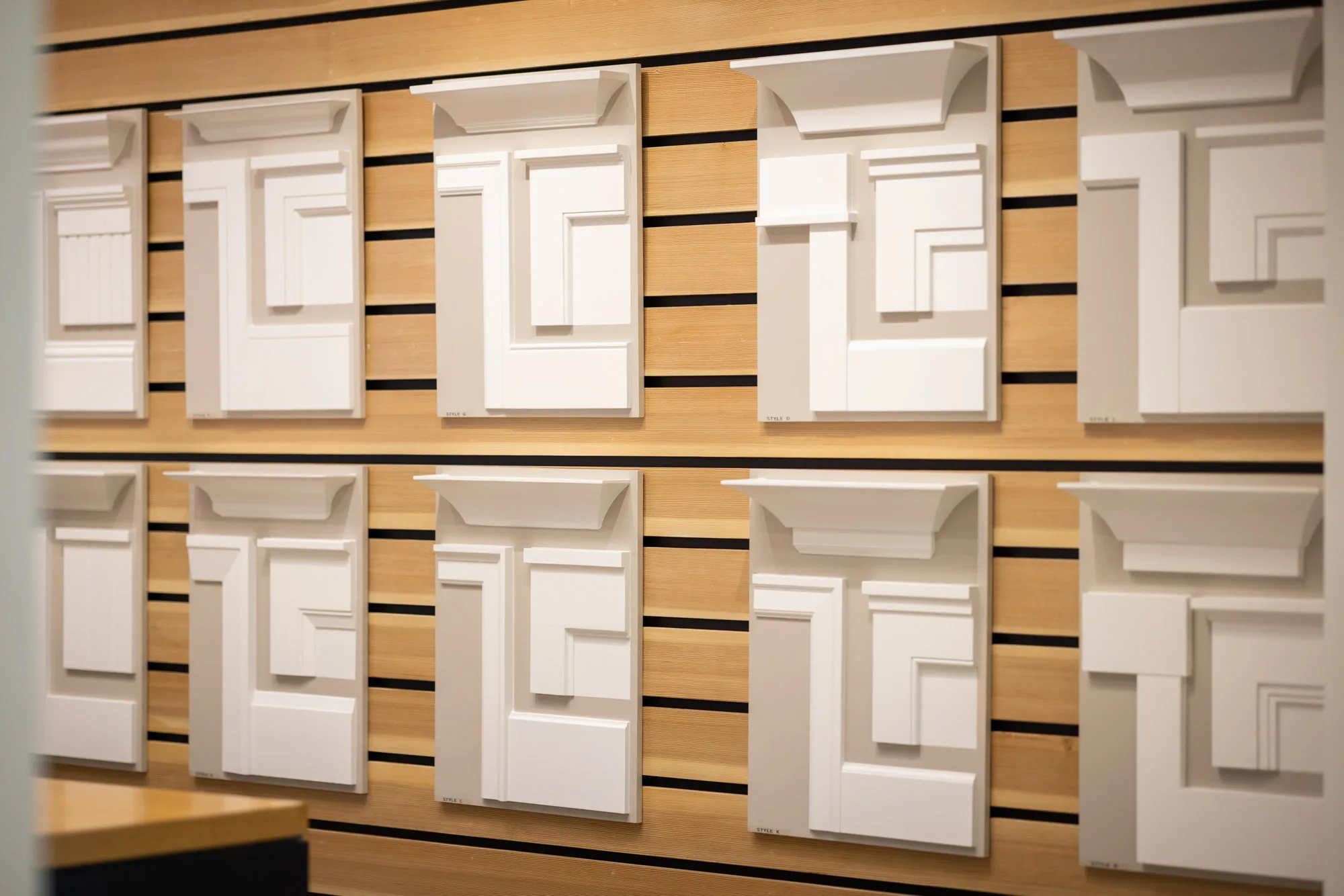 Wall display of white decorative molding corner samples mounted on wooden slats, arranged in two rows with different styles and shapes for interior design.