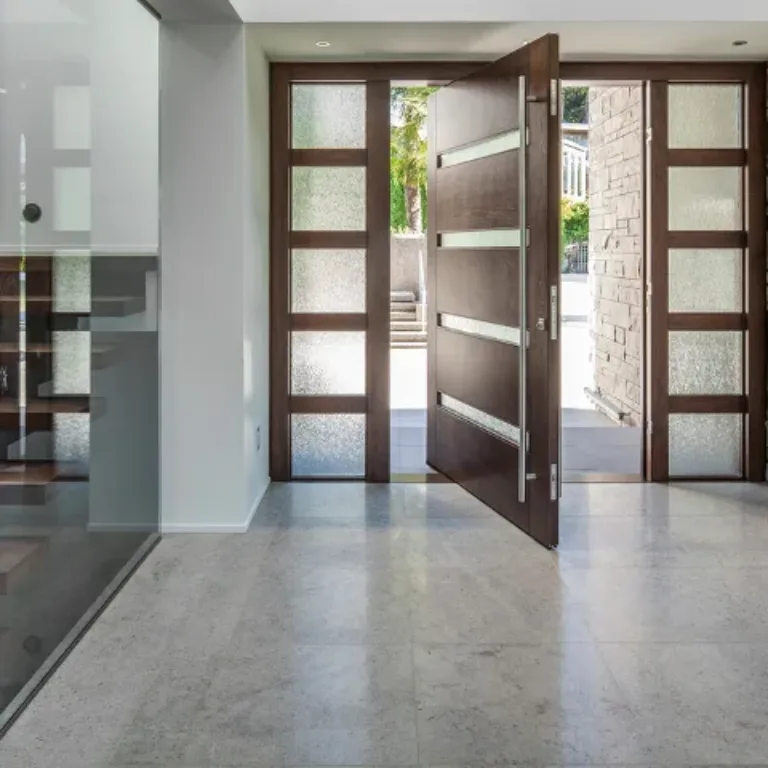 Modern interior entryway with an open dark wooden front door featuring horizontal frosted glass panels, flanked by glass-paneled sidelights, light gray polished stone floor, stone wall on the right, and a staircase with wooden steps and glass railing on the left.