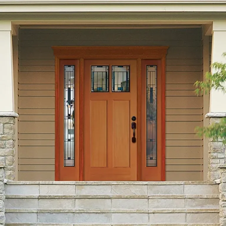 Front entrance of a house featuring a wooden door with decorative glass panels on each side and above, framed by beige siding, stone pillars, white railings, and surrounding greenery.