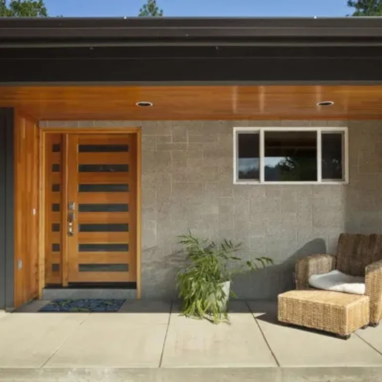 Modern house entrance with a wooden front door featuring horizontal glass panels, concrete block wall, a small rectangular window, a potted green plant, and a wicker chair with a white cushion on a concrete porch.