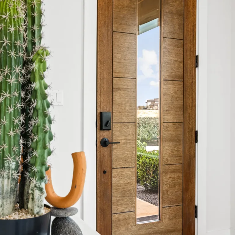 Modern wooden front door with a vertical glass panel, showing a view of an outdoor pathway, green bushes, and partly cloudy sky, with a large potted cactus and decorative object in the foreground.
