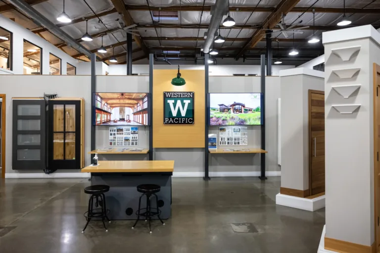 Modern showroom interior with polished concrete floor, two black stools at a central wooden desk, wall displays featuring wooden doors and two large digital screens showing house interiors and exteriors, and a prominent Western Pacific sign in the center under a green industrial-style lamp.