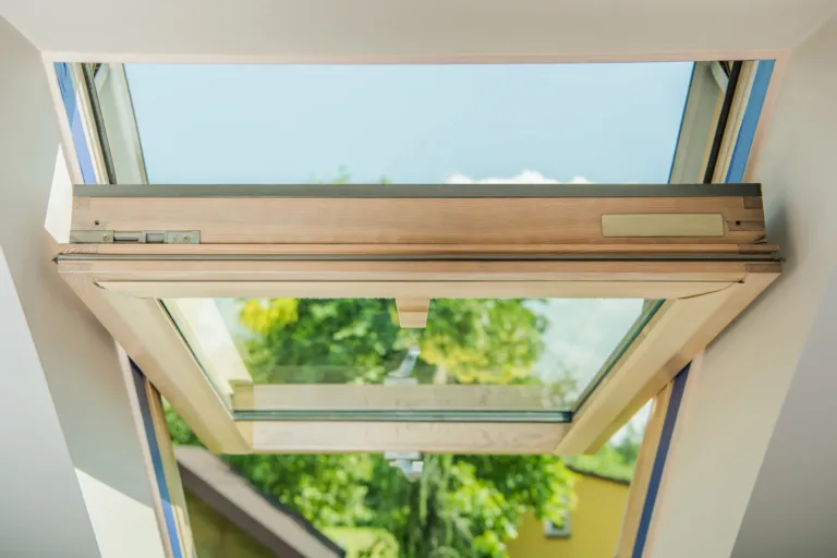 Open skylight window with wooden frame showing clear blue sky and green trees outside, viewed from inside a building.