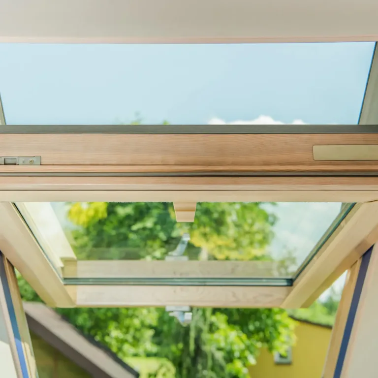 Open skylight window with wooden frame showing clear blue sky and green trees outside, viewed from inside a building.