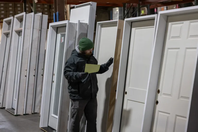 A person wearing a green beanie, black jacket, and gloves is inspecting a white door in a warehouse filled with multiple white doors standing upright.