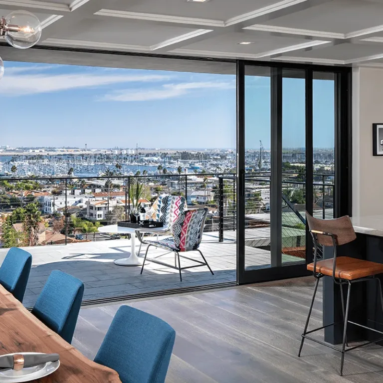 Modern open-plan kitchen and dining area with blue chairs around a wooden dining table set with plates and napkins, a kitchen island with brown stools, geometric patterned backsplash, and large sliding glass doors opening to a balcony overlooking a marina and cityscape under a blue sky.