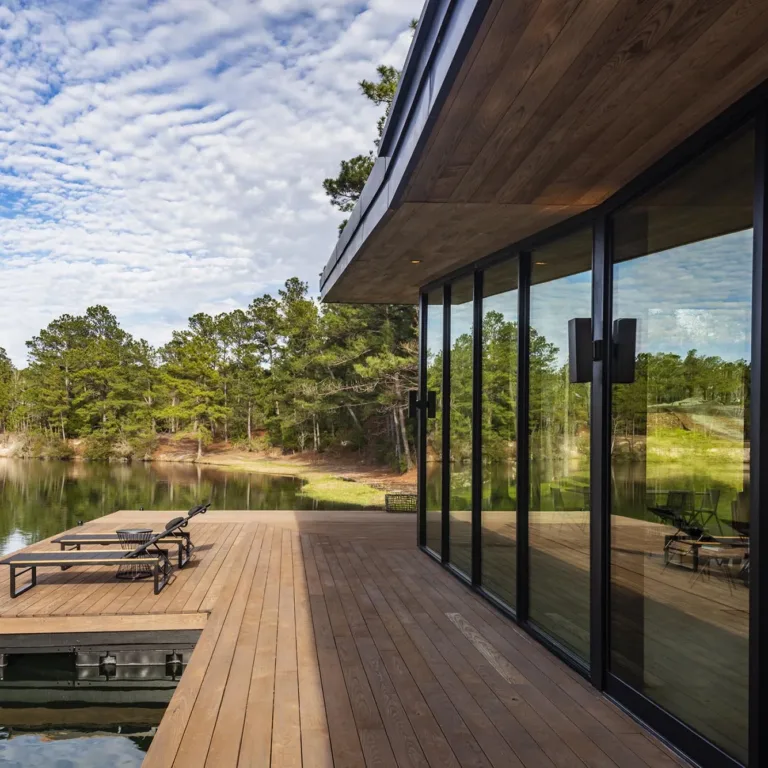 Wooden deck extending over a calm lake with two lounge chairs and a basket, adjacent to a modern building with large glass windows reflecting trees and sky.
