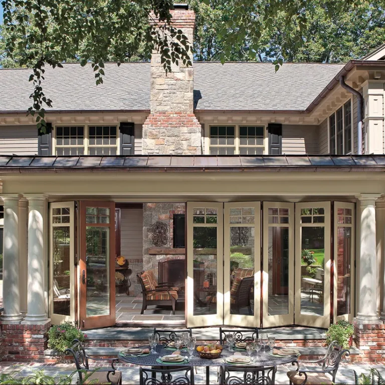 Backyard patio of a two-story traditional house with beige siding, multiple windows, a stone chimney, and large glass doors opening to an outdoor seating area with a round metal table and black wrought iron chairs, surrounded by brick columns and greenery.