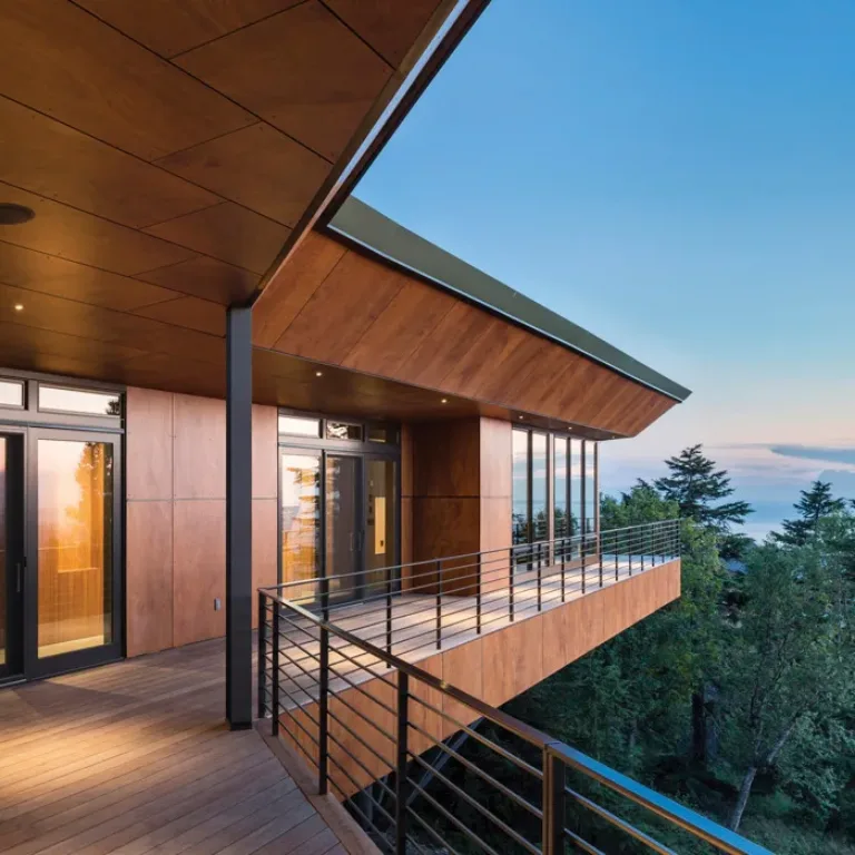 Modern wooden balcony with glass doors and metal railing overlooking tall trees and a clear blue sky at dusk