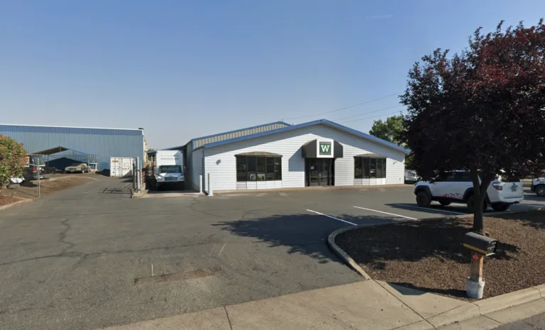 Single-story white commercial building with a Western Pacific sign above the entrance, adjacent parking lot with a white SUV, a mailbox on a wooden post, and a tree with dark red leaves under a clear blue sky.