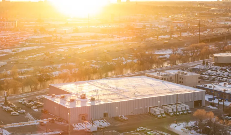 Large industrial warehouse with multiple loading dock trucks parked outside, adjacent to a busy highway at sunset with sunlight creating a golden glow over the snow-covered landscape and bare trees.