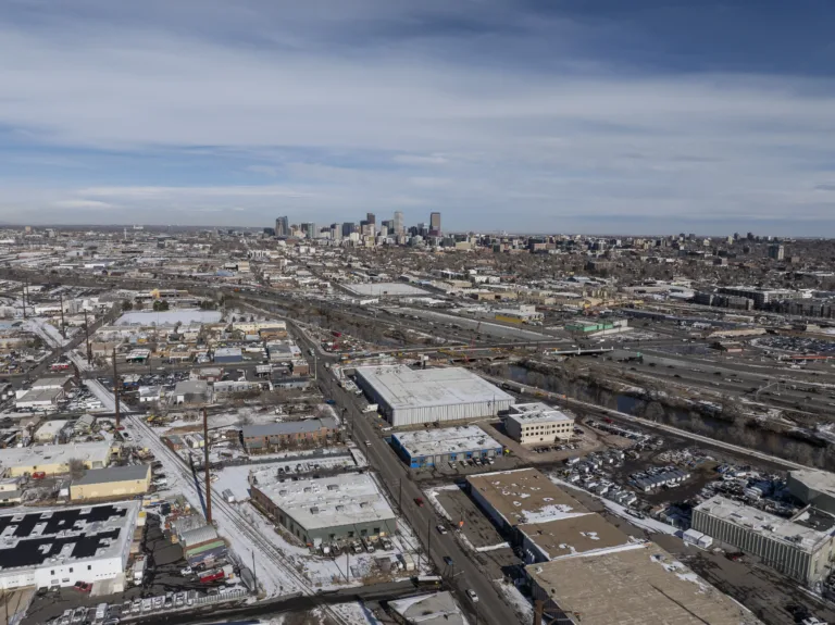Aerial view of an industrial area with warehouses and roads covered in light snow, with a highway and a city skyline visible in the distance under a partly cloudy sky.