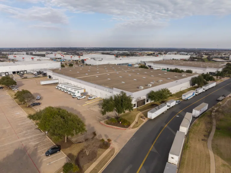 Aerial view of a large industrial warehouse complex with multiple trucks and trailers parked along the building and road, surrounded by parking lots, trees, and a partly cloudy sky.