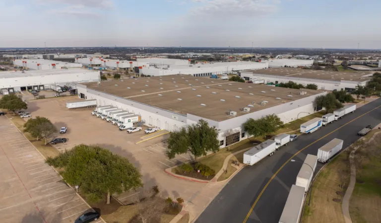 Aerial view of a large industrial warehouse complex with multiple trucks and trailers parked along the building and road, surrounded by parking lots, trees, and a partly cloudy sky.