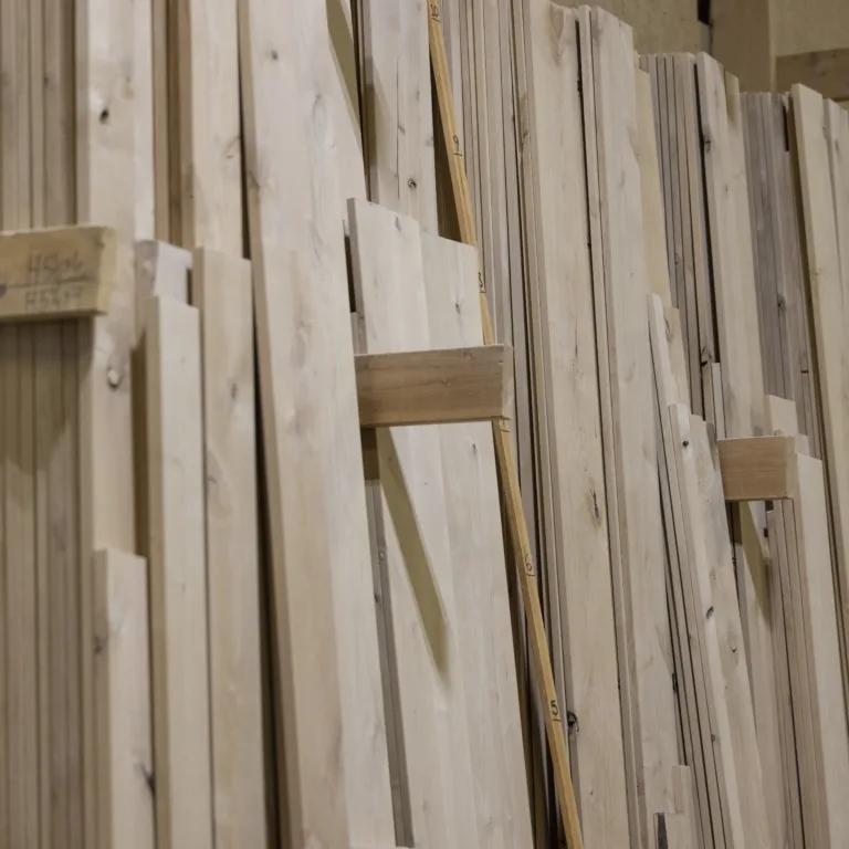 Stacked light-colored wooden planks and boards neatly arranged vertically in a woodworking or lumber storage area.