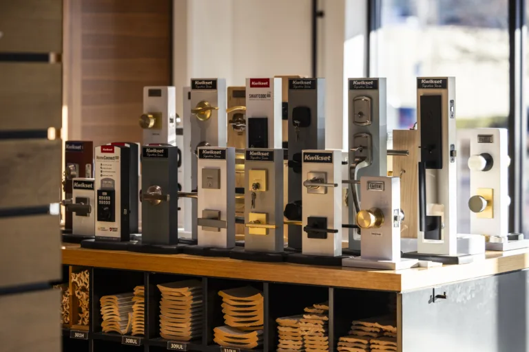 Display of various door lock models mounted on wooden stands inside a store, featuring brands like Kwikset and Emtek with different handle designs, levers, knobs, and finishes, arranged on a wooden countertop with shelves underneath holding door hardware parts.