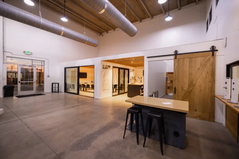 Spacious modern industrial office interior with polished concrete floor, exposed ductwork on high ceiling, a small table with two black stools in the foreground, glass-walled conference rooms with warm lighting in the background, and a large sliding wooden barn door on the right side.