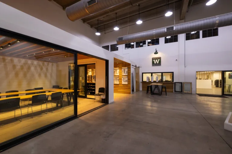 Indoor modern office space with large glass-walled conference room on the left, a wooden table and black chairs inside, polished concrete floor, exposed ceiling with ductwork and hanging lights, and a reception or display area with a Western Pacific sign on the far wall and additional office spaces beyond.