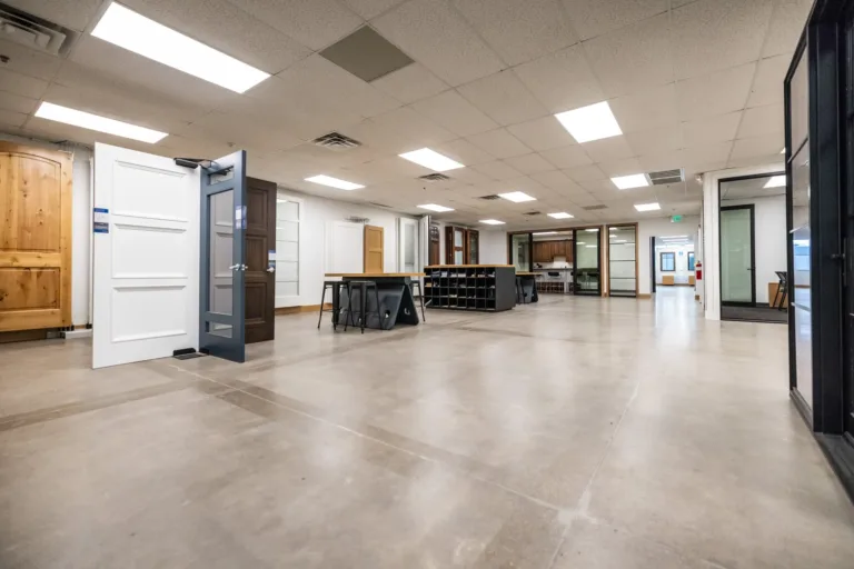 Interior showroom featuring multiple display doors in various colors and styles standing upright on a polished concrete floor under bright ceiling lights in a spacious setting with tables and stools in the background.