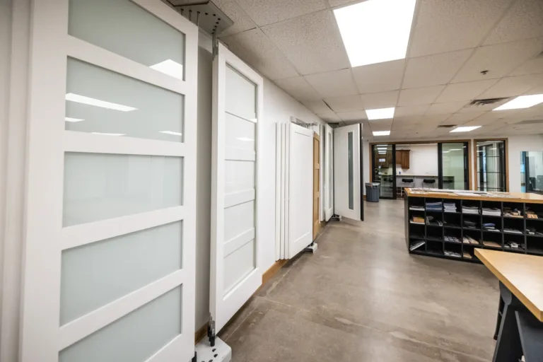 Interior showroom displaying multiple modern sliding glass doors with white frames on the left, an open office space with concrete floor, fluorescent ceiling lights, and cubby shelves filled with papers and materials on the right.