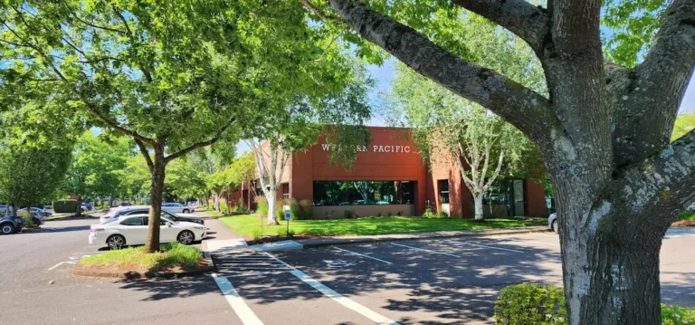 Wide parking lot with several parked cars and green trees surrounding a brick office building with a sign that reads Western Pacific, under a bright blue sky.