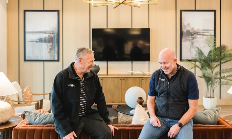 Two men sitting on the back of a brown sofa in a modern living room, smiling and talking, with a large TV and wooden cabinet in the background, along with framed pictures and a potted plant.