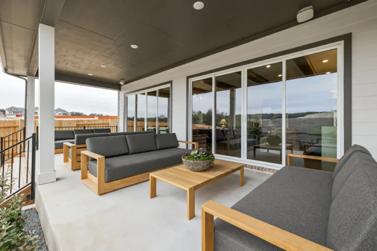 Covered outdoor patio with three gray cushioned wooden sofas arranged around a low wooden coffee table with a potted succulent centerpiece, sliding glass doors reflecting the outdoor surroundings, and a fenced backyard with houses in the distance.