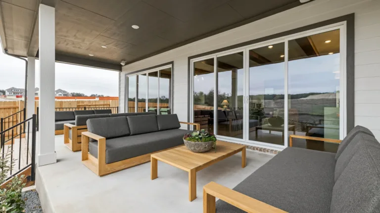 Covered outdoor patio with three gray cushioned wooden sofas arranged around a low wooden coffee table with a potted succulent centerpiece, sliding glass doors reflecting the outdoor surroundings, and a fenced backyard with houses in the distance.