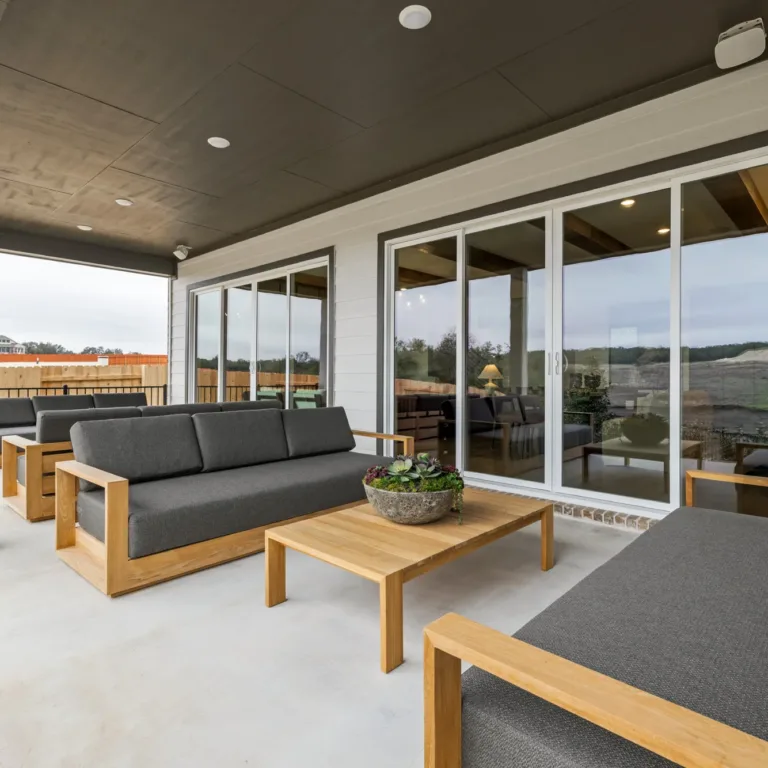 Covered outdoor patio with three gray cushioned wooden sofas arranged around a low wooden coffee table with a potted succulent centerpiece, sliding glass doors reflecting the outdoor surroundings, and a fenced backyard with houses in the distance.