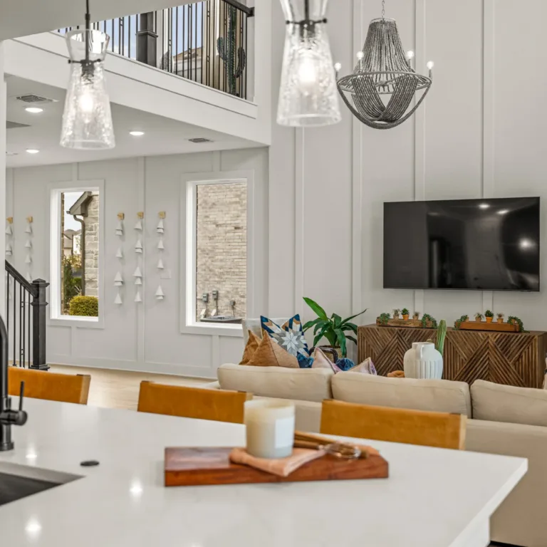 Modern open-concept living space featuring a kitchen island with a black faucet and light wood cabinets, hanging pendant lights above, a beige sectional sofa with colorful pillows, a wall-mounted flat-screen TV, wooden furniture with plants, and framed artwork on the wall in the background.