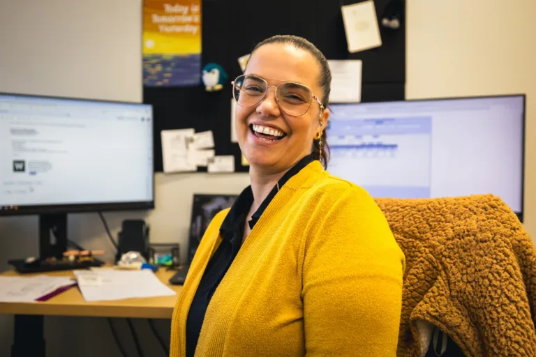 Smiling woman wearing glasses and a yellow sweater sitting at a desk in an office with two computer monitors and papers in the background.