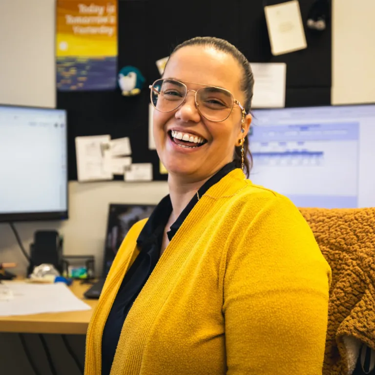 Smiling woman wearing glasses and a yellow sweater sitting at a desk in an office with two computer monitors and papers in the background.