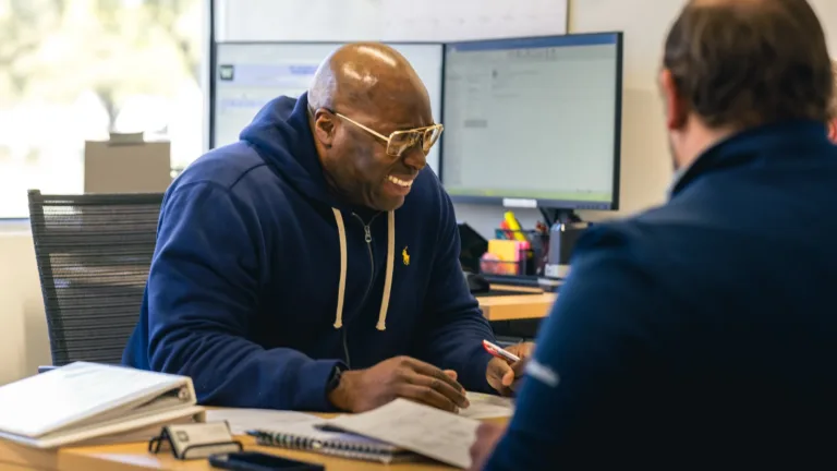 Two men sitting at a desk in an office, one man wearing a navy hoodie and glasses is smiling and holding a pen, the other man is seen from behind wearing a dark blue shirt, with papers, a notebook, and a smartphone on the desk and computer monitors in the background.