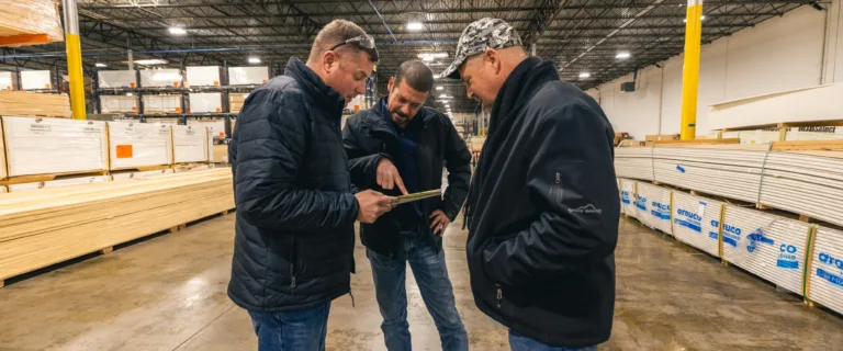 Three men in jackets standing and discussing something while looking at a tablet inside a warehouse with stacks of lumber on shelves and the floor around them.