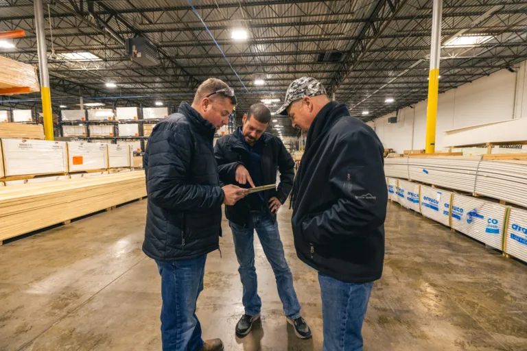 Three men in jackets standing and discussing something while looking at a tablet inside a warehouse with stacks of lumber on shelves and the floor around them.