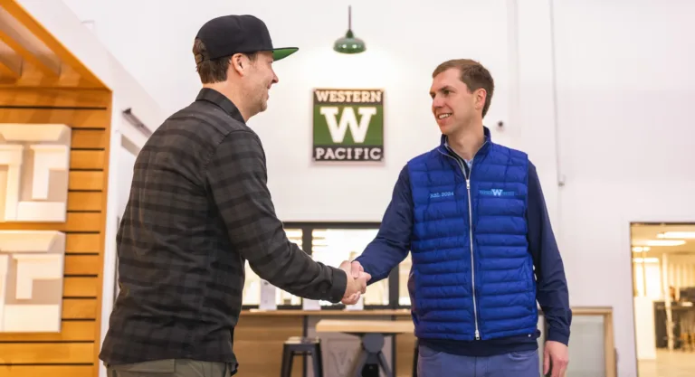 Two men shaking hands indoors in front of a sign that reads Western Pacific, one wearing a black cap and checkered shirt, the other wearing a blue sleeveless jacket and navy long-sleeve shirt.