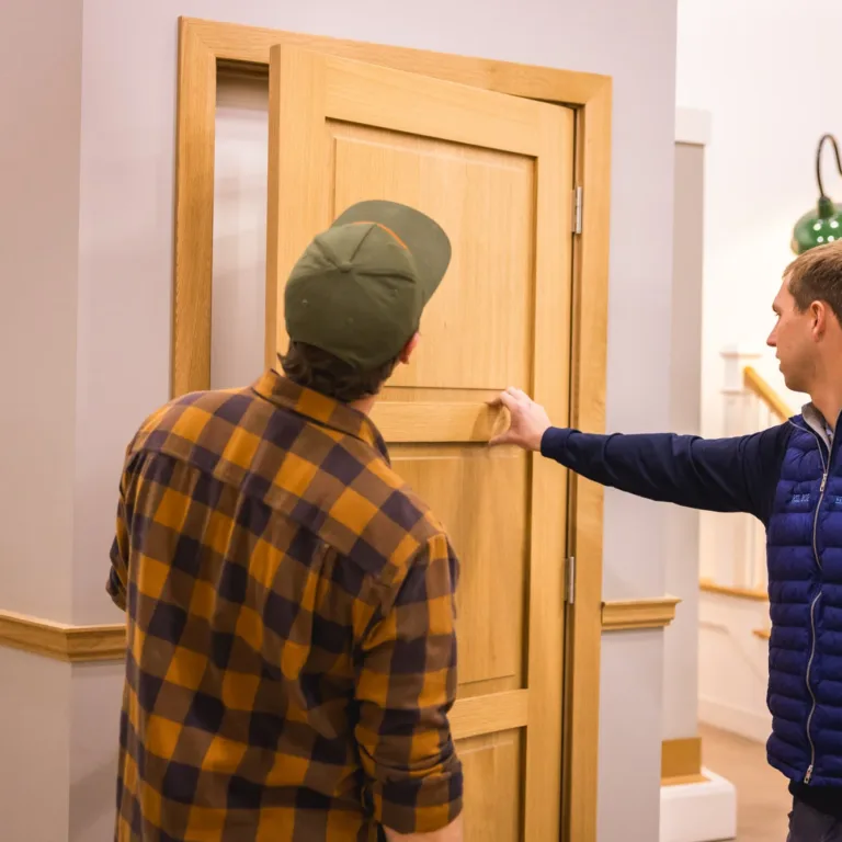 Two men examining a wooden door indoors, one wearing a green cap and checkered shirt, the other in a blue vest pointing at the door's surface.