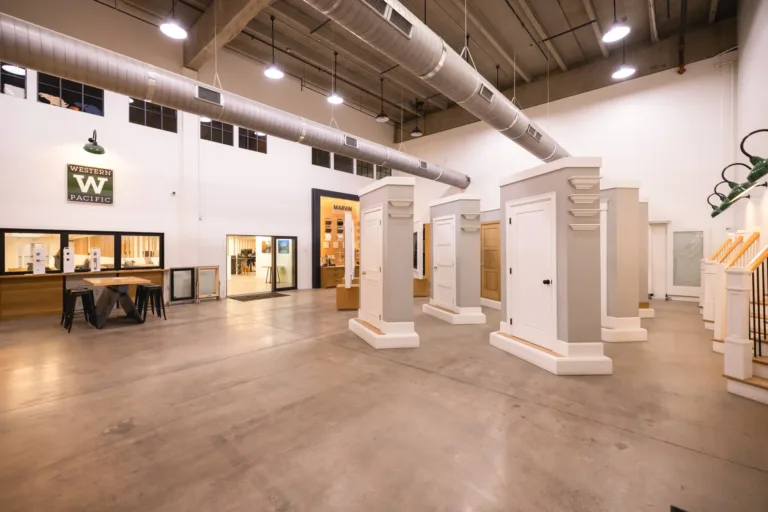 Spacious showroom with high ceilings displaying various white and wood-paneled door models and stair railing samples under industrial-style lighting and exposed ductwork.