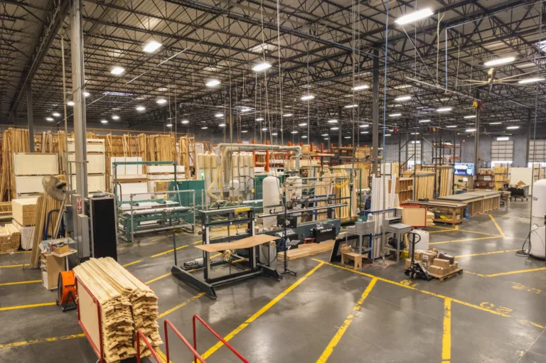 Interior of a large woodworking factory with stacks of wooden planks, various woodworking machines, equipment, and organized storage racks under a high ceiling with industrial lighting.