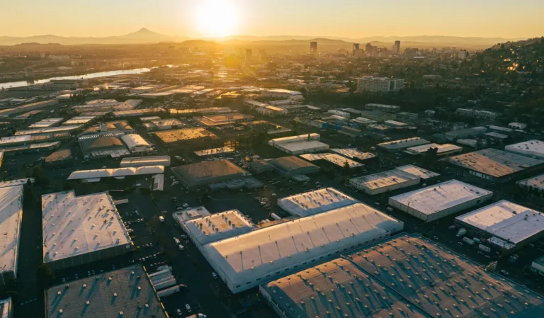 Aerial view of an industrial area with numerous warehouses and buildings under the golden light of a setting sun, a river and distant mountains are visible on the horizon.