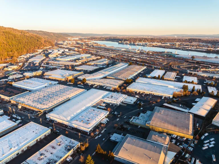 Aerial view of an industrial area with numerous warehouses featuring white and gray roofs, adjacent to a river with cranes and shipping docks, and surrounded by forested hills under a clear sky at sunset.