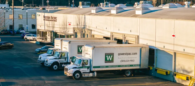 Three white Western Pacific delivery trucks parked in loading bays outside a large warehouse building with numbered bays, surrounded by parked cars and trees on a hillside in the background.