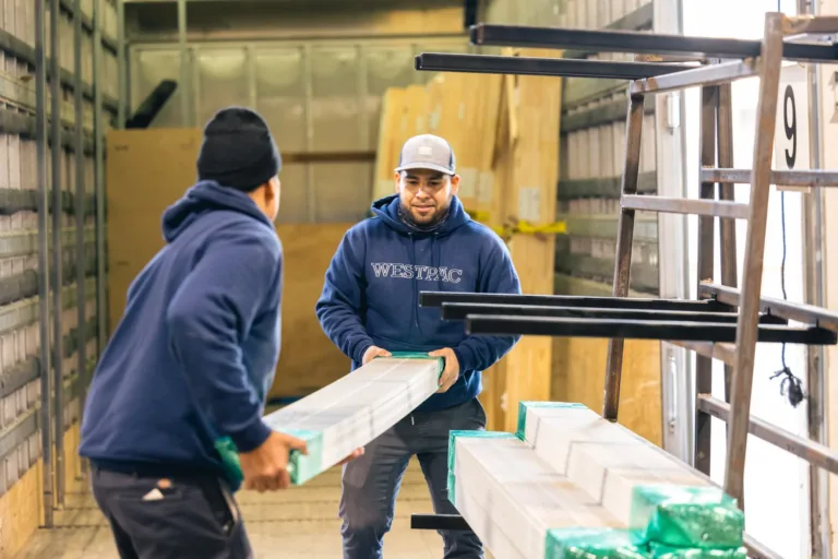 Two men inside a truck unloading long, wrapped materials, with one man wearing a WESTPAC hoodie and a cap, and the other wearing a beanie and sweatshirt.