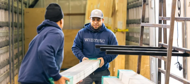 Two men inside a truck unloading long, wrapped materials, with one man wearing a WESTPAC hoodie and a cap, and the other wearing a beanie and sweatshirt.
