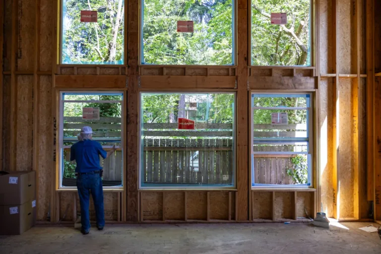 A person installing or inspecting large rectangular windows in a wooden framed wall of a building under construction, with a view of trees and a wooden fence outside.
