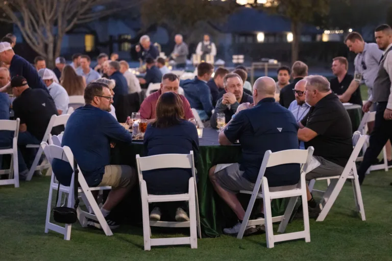 Group of people seated around a round table outdoors at dusk, engaged in conversation with drinks and food on the table, with more attendees in the background and a waiter walking nearby.