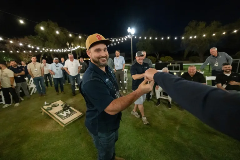 A man wearing a tan St. Louis Cardinals cap and navy shirt smiling and fist-bumping another person at an outdoor night event with string lights and a crowd of people and a cornhole board in the background.